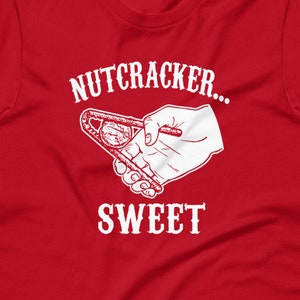May include: A red t-shirt featuring the text "NUTCRACKER... SWEET" in white, accompanied by a graphic of a hand using a nutcracker. The design is printed on the front of the shirt.