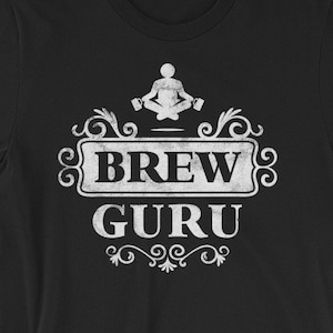 Può includere: Maglietta nera con una grafica bianca di una persona che medita con due tazze nelle mani. La grafica è circondata da una cornice decorativa e dal testo "Brew Guru".