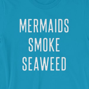 May include: A turquoise t-shirt with the white text "MERMAIDS SMOKE SEAWEED" in a stacked layout. The shirt is a short-sleeved crew neck style.