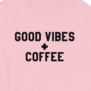 Może przedstawiać: Różowa koszulka z krótkim rękawem z czarnym napisem "GOOD VIBES + COFFEE".