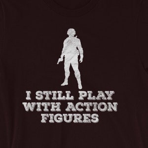 Könnte beinhalten: Ein dunkelmarrones T-Shirt mit einer weißen Silhouette einer Person, die eine Waffe hält, und dem Text "I still play with action figures".