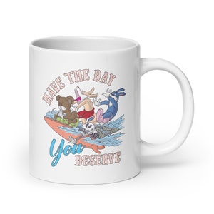 Könnte beinhalten: Weiße Keramiktasse mit einer Cartoon-Illustration eines Kaninchens, eines Bären, eines Skeletts und eines blauen Hasen, die auf einem Surfbrett reiten. Der Text "HAVE THE DAY YOU DESERVE" ist auf die Tasse gedruckt.
