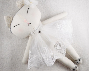 May include: A white fabric cat doll wearing a white tulle skirt with lace trim. The doll has a white flower on its head and silver glitter on its ears.