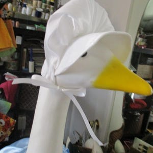 May include: A white goose sculpture wearing a white bonnet and a light blue dress. The goose has a yellow beak and a white ribbon tied around its neck. The sculpture is a whimsical decorative item.