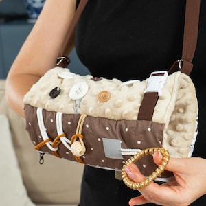 Op de afbeelding: Een beige en bruine sensorische tas met verschillende texturen en sluitingen, waaronder knopen, een gesp en een rits. De tas heeft een bruine riem en is ontworpen voor tactiele exploratie. Een goudkleurige armband wordt in de hand gehouden.