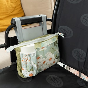 Może przedstawiać: Boczna torba do wózka inwalidzkiego z kwiatowym nadrukiem stokrotek. Posiada główną komorę zapinaną na zamek i mniejszą kieszeń. Torba ma jasnoniebieskie tło ze wzorem stokrotek i zieloną kieszeń akcentującą. Jest przymocowana do podłokietnika szarymi paskami.