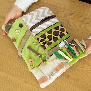 Puede incluir: Un tablero de actividades sensoriales con varias texturas y elementos interactivos. El tablero presenta una combinación de colores verde y beige con botones, cremalleras, cintas y otros componentes táctiles. Diseñado para estimular la exploración sensorial y la motricidad fina.