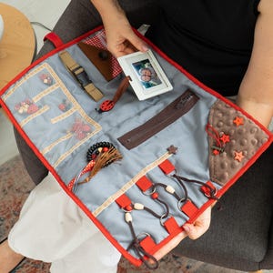 Puede incluir: Un tablero de actividades sensoriales con varias texturas y elementos interactivos. Cuenta con una base de tela gris con ribete rojo, una cremallera marrón, una hebilla, una mariquita y un pequeño marco de fotos. Incluye cuentas de madera y botones en forma de estrella.