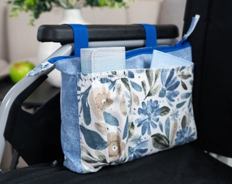 May include: A blue and white floral patterned bag with blue straps hangs on the back of a wheelchair. The bag has multiple pockets and is designed to hold personal items.