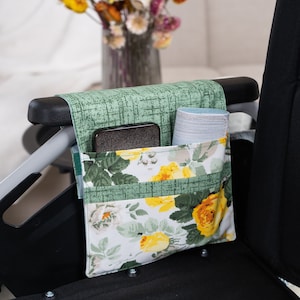 May include: A green and white floral fabric organizer with pockets for a phone and other items. The organizer is attached to the armrest of a wheelchair.