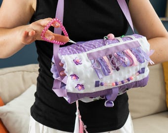 May include: A sensory activity roll featuring a white dimpled fabric base with purple accents. The roll includes various tactile elements such as colorful ribbons, plastic beads, flower-shaped embellishments, and a white and pink tassel hanging from the bottom.