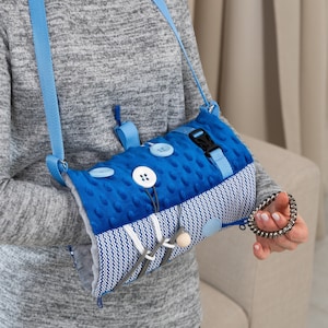 Puede incluir: Un juguete sensorial azul y blanco con botones, cremalleras y una cuenta de madera. El juguete se lleva en el brazo con una correa azul.