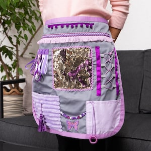 Peut inclure: Tablier sensoriel gris et violet, orné de diverses textures et décorations. Il comprend un carré à paillettes, des rubans, un pompon et un papillon perlé. Le tablier est conçu avec plusieurs poches et éléments décoratifs.