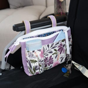 May include: A lavender and white floral storage pouch with two top straps for attachment. The pouch features a zippered main compartment, two front pockets, and a white strap with a metal clip holding a set of keys with a blue circular tag.