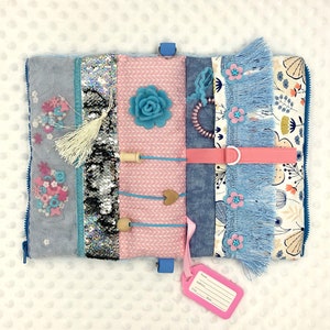 Op de afbeelding: Een blauw en roze baby-activiteitenboek met verschillende texturen en functies, waaronder een rits, knopen, een hart, een bloem, een kwast en een label met de woorden "Naam", "Adres", "Stad" en "Staat".