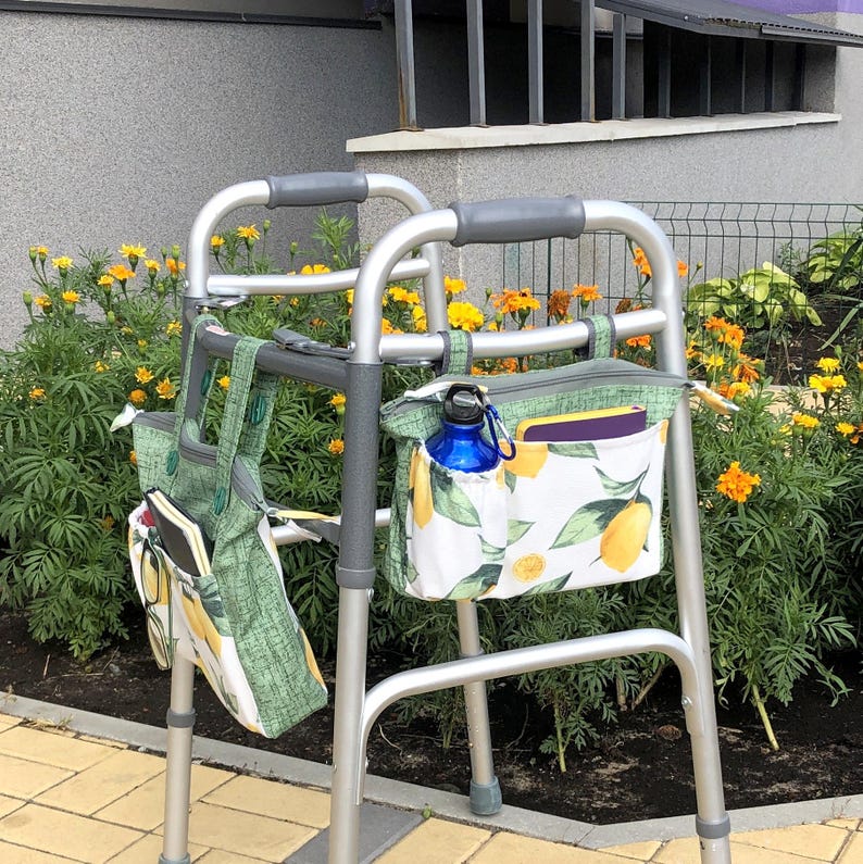 Op de afbeelding: Een zilverkleurige metalen rollator met twee bevestigde stoffen opbergtassen met een patroon van gele citroenen en groene bladeren op een witte achtergrond. De tassen hebben zijvakken met een blauwe waterfles en een paars notitieboekje.