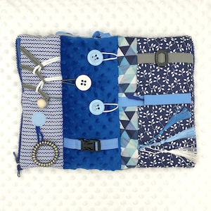 Puede incluir: Un tablero de actividades sensoriales con varias texturas y cierres. Cuenta con una cremallera azul, botones, hebillas y telas texturizadas en tonos azules y blancos. Incluye una cuenta de madera y una espiral metálica.