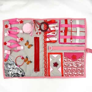 Puede incluir: Un tablero de actividades sensoriales con varias texturas y elementos. Cuenta con cintas rosas, una cremallera roja, un pompón rosa esponjoso y detalles de mariposas. Incluye un bolsillo con un estampado floral y una flor roja.