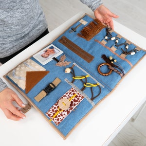 Può includere: Un tappetino di attività in denim blu con diverse texture e oggetti per il gioco sensoriale. Presenta una foto, una cerniera, una fibbia, un fiocco marrone, un anello di legno e un tessuto con motivo marrone e bianco.