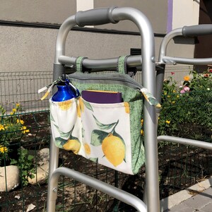 Op de afbeelding: Een zilverkleurige metalen rollator met een stoffen opbergtas bevestigd aan het voorframe. De tas heeft een patroon van gele citroenen en groene bladeren op een witte achtergrond, een zijvak met een blauwe waterfles en een paars notitieboek.