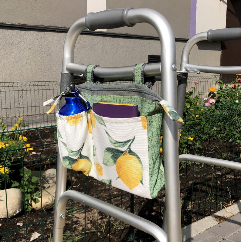 Op de afbeelding: Een zilverkleurige metalen rollator met een stoffen opbergtas bevestigd aan het voorframe. De tas heeft een patroon van gele citroenen en groene bladeren op een witte achtergrond, een zijvak met een blauwe waterfles en een paars notitieboek.