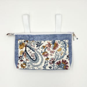 Może przedstawiać: Niebieski, materiałowy organizer do zawieszenia z kieszenią we wzory kwiatowe. Kieszeń ma wzór paisley w odcieniach różu, żółci i niebieskiego. Białe paski są przymocowane u góry do zawieszenia. Zamek błyskawiczny biegnie wzdłuż górnej krawędzi.