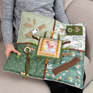 Puede incluir: Una alfombra de actividad de tela verde y marrón con varias texturas y elementos para bebés y niños pequeños. Cuenta con un marco de fotos, una cremallera, una hebilla, un anillo de madera, una borla, botones y hojas.