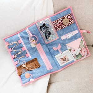 Peut inclure: Un tableau d'activités sensorielles avec une base en tissu bleu imitation jean et une bordure rose. Comprend une fermeture éclair, une boucle, un bouton, un cadre photo et diverses textures et formes pour l'exploration tactile.