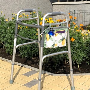Op de afbeelding: Een zilverkleurige metalen rollator met grijze rubberen handgrepen en een stoffen opbergtas bevestigd aan het voorframe. De tas heeft een witte achtergrond met een patroon van gele citroenen en groene bladeren, met een blauwe waterfles erin.