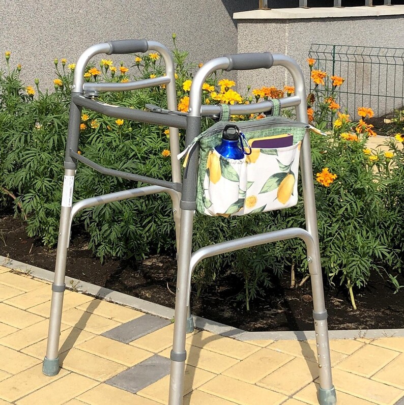 Op de afbeelding: Een zilverkleurige metalen rollator met grijze rubberen handgrepen en een stoffen opbergtas bevestigd aan het voorframe. De tas heeft een witte achtergrond met een patroon van gele citroenen en groene bladeren, met een blauwe waterfles erin.