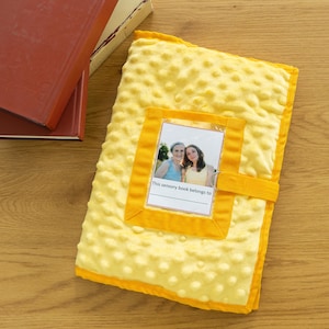 Puede incluir: Un libro sensorial amarillo con una superficie texturizada y punteada. Cuenta con un bolsillo transparente con una foto y el texto "This sensory book belongs to". Una correa amarilla asegura el libro. Dos libros son visibles en el fondo.