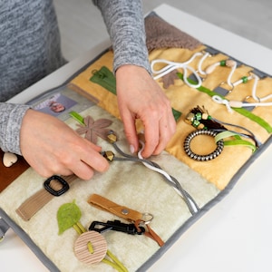 May include: A sensory activity board with various textures and interactive elements. Features include a buckle, zipper, button, and a keychain. The board is made of soft, colourful fabrics, including brown, yellow, and green, designed for tactile exploration and fine motor skill development.