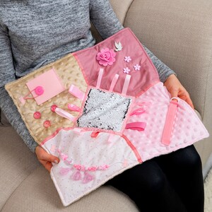 May include: A pink and beige baby sensory activity mat with various textures, including sequins, ribbons, and buttons. The mat has a loop for attaching a toy.