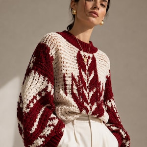 May include: A burgundy and cream-colored crochet sweater with a leaf-like pattern. The sweater has a crew neck and long sleeves. The model is wearing white pants and gold earrings.