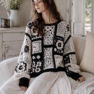 Peut inclure: Un pull en crochet crème et noir avec un motif floral. Le pull a un col rond et des manches longues avec des poignets noirs. Le design incorpore des motifs carrés et floraux. La personne porte une jupe blanche.