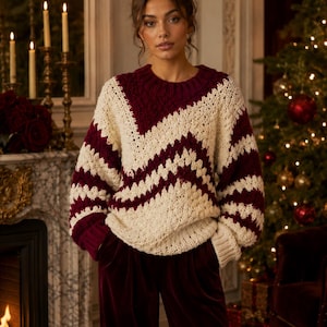 May include: A cozy, oversized sweater in cream and burgundy with a chevron pattern. The sweater has a crew neck and long sleeves. The model is wearing burgundy velvet pants. A Christmas tree and fireplace are in the background.