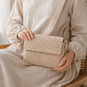 May include: A beige rectangular crossbody bag crafted from woven material with a flap closure and a matching shoulder strap. The bag features a consistent horizontal ribbed texture throughout its exterior surface, providing a structured appearance.