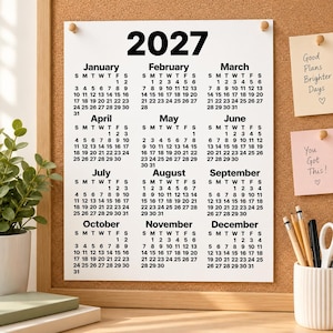 Minimalist 2027 one-page calendar pinned to a corkboard with wooden pins, styled with pastel notes, a potted plant, stacked books and stationery cup in a warm organised desk space.