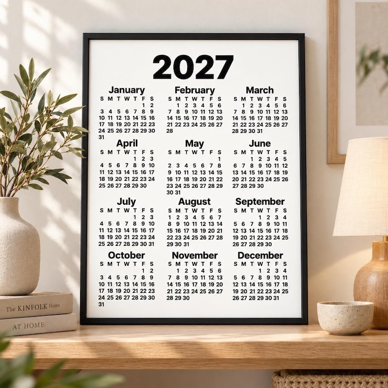 Minimalist 2027 printable calendar in a black frame displayed on a wooden shelf, surrounded by a vase with greenery, stacked books, table lamp and ceramic bowl in a soft Scandinavian-style interior.