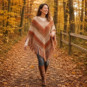 May include: A crochet poncho featuring a V-neckline and a chevron pattern in shades of cream, tan, and burnt orange. The garment has a loose, triangular silhouette with long, dangling fringe trim along the bottom hem, worn over a white shirt.