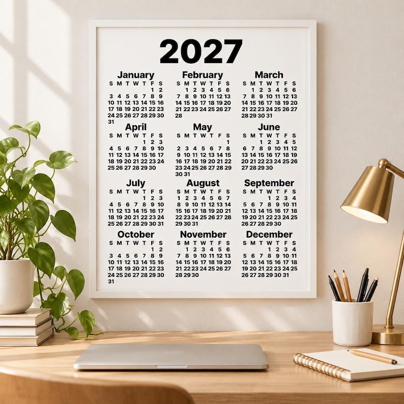 Minimalist 2027 one-page black-and-white calendar in a white frame hanging above a wooden desk, styled with a green plant, brass lamp, laptop, pencils and notebook in a bright neutral home office.