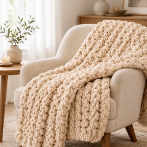 May include: A chunky knit throw blanket in a solid cream color, featuring a thick, textured yarn construction with a repeating vertical stitch pattern that drapes over the seat and back of an upholstered armchair with wooden legs.
