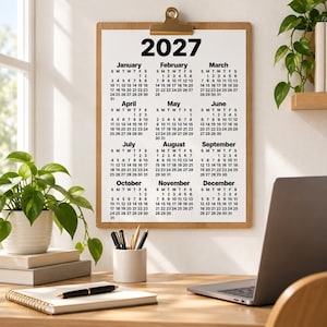 Minimalist 2027 yearly calendar clipped to a wooden clipboard on a sunlit wall above a desk, with a laptop, notebook, pen holder and green plants creating a clean modern workspace scene.