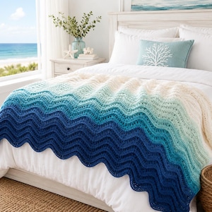 May include: A crochet blanket featuring a wavy ripple pattern with an ombre color gradient transitioning from deep navy blue at the bottom to light aqua and white at the top, draped over a white bedspread with a matching teal coral-print pillow.