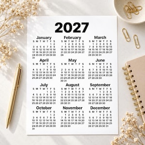 Minimalist 2027 printable calendar shown as a flat lay on a light desk surface, styled with dried flowers, a gold pen, paper clips, ceramic dish and spiral notebook for an elegant stationery-themed mockup.