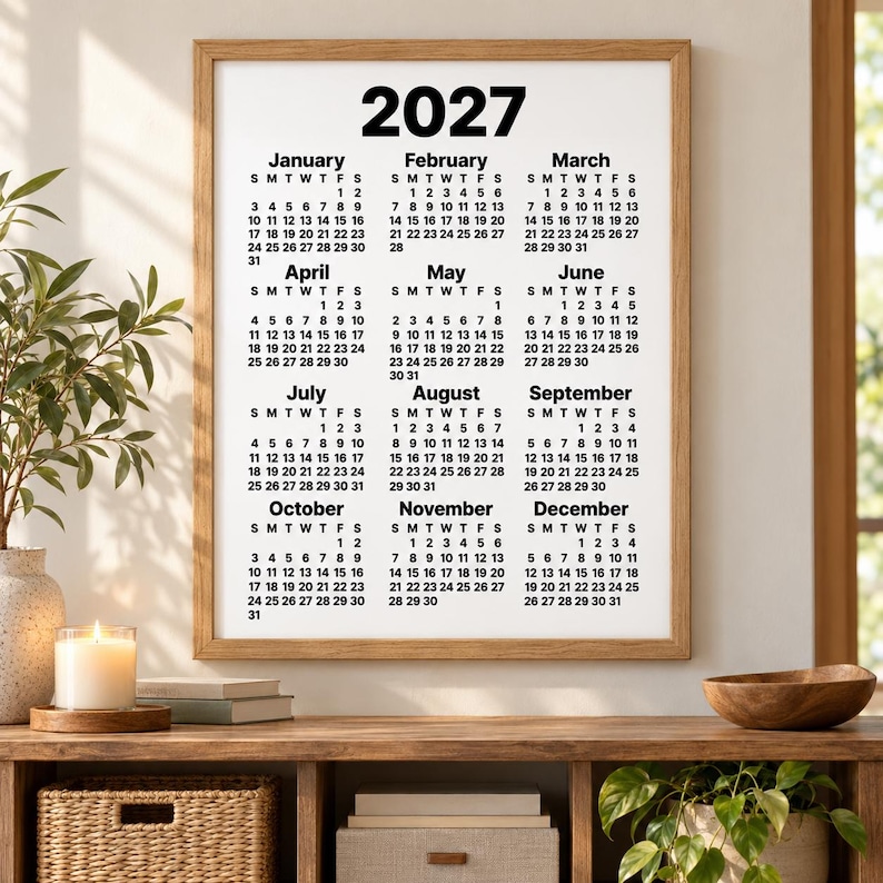 Minimalist 2027 black-and-white wall calendar in a natural oak frame above a wooden console table, styled with a candle, books, woven basket, decorative bowl and potted plants in a cosy home setting.