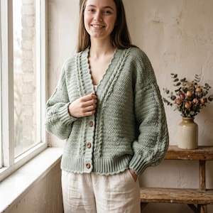 May include: A light green, hand-crocheted cardigan with a scalloped edge and wooden buttons. The sweater features a cable knit design on the front and sleeves. The cardigan is paired with beige pants. The overall style is casual and comfortable.