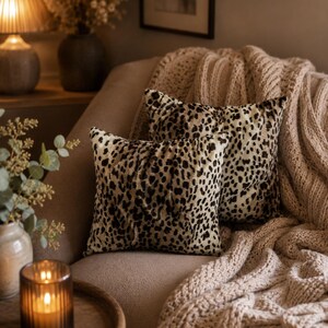 May include: Two square throw pillows with a leopard print pattern rest on a beige sofa, complemented by a chunky knit blanket. The pillows feature a tan background with dark brown spots, adding a touch of animal print to the room's decor.