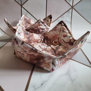 Op de afbeelding: Twee bruine en witte stoffen kom-warmers met een bloemen- en dierenprint. De warmers zijn gevouwen en op elkaar gestapeld.