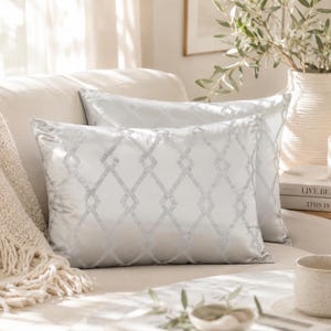 May include: Two rectangular, light grey throw cushions with a geometric trellis pattern on a cream sofa. A beige throw blanket with fringe is draped on the sofa. A vase with greenery and a lit candle are visible.